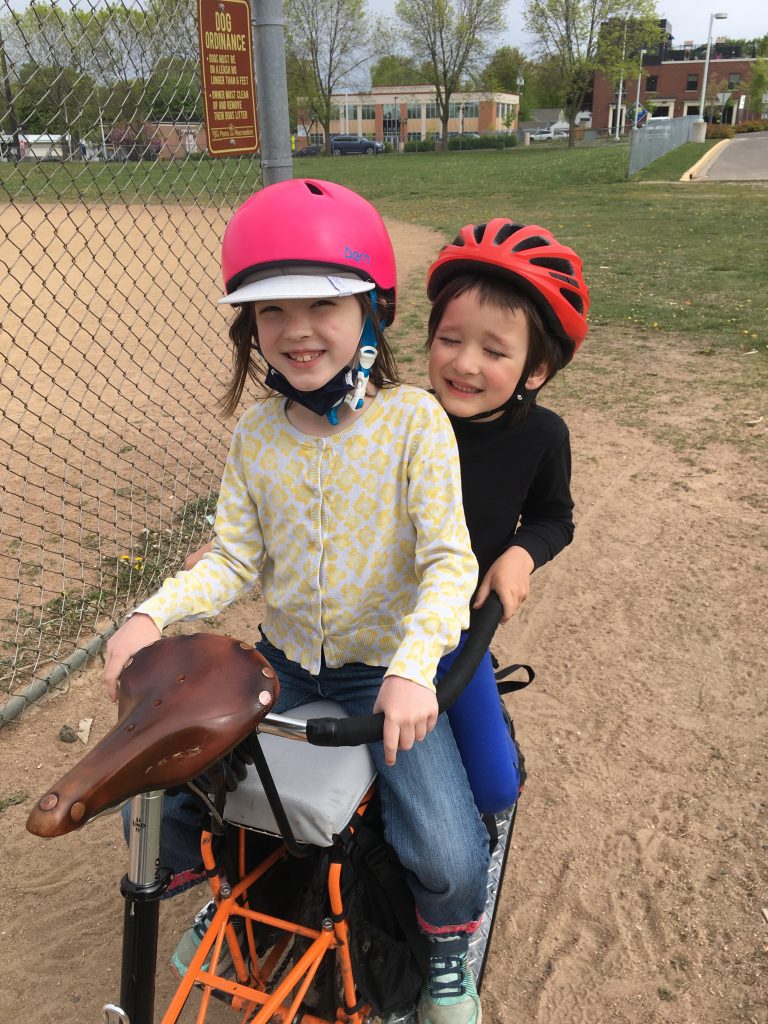 Two children sitting on a cargo bike passenger attachment on a sandy surface.