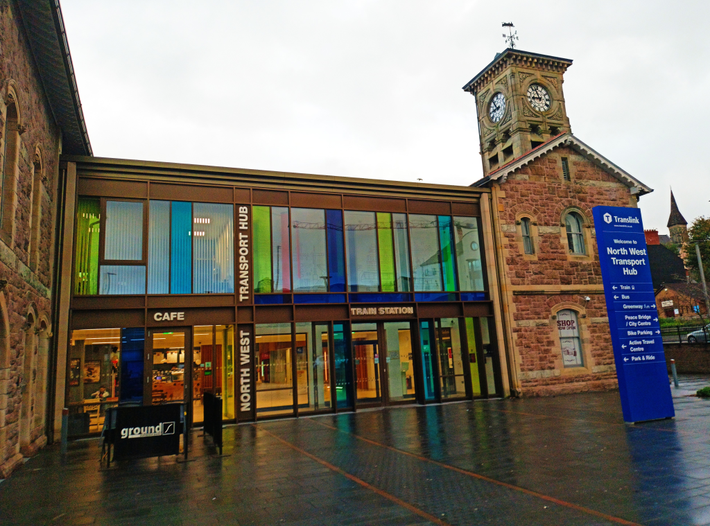 The railway station in Derry, Ireland, has modern floor-to-ceiling windows set inside a stone structure with a clock tower.