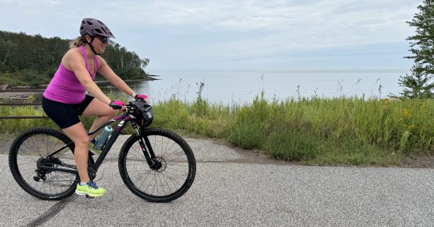 A woman rides a road bike on a paved trail with Lake Superior to her left.