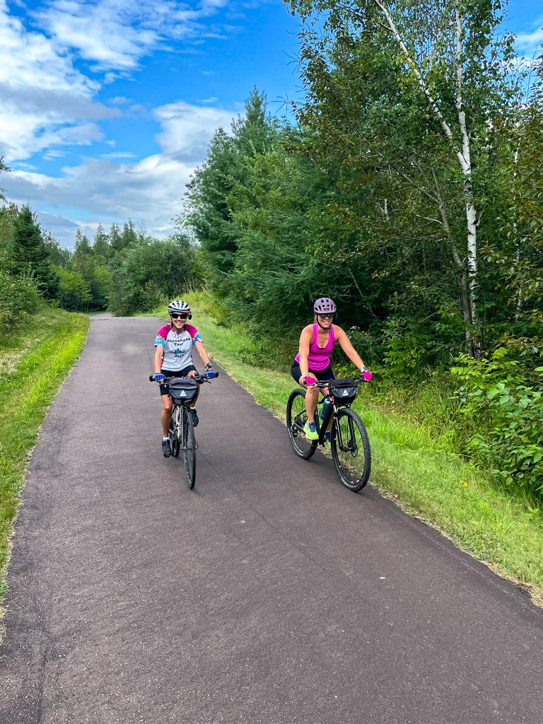 Two cyclists along a paved trail through a wooded area. 
