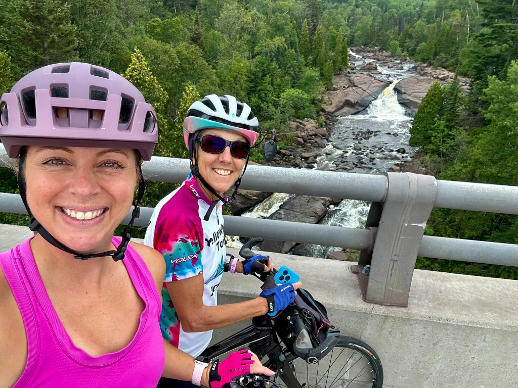 Two cyclists pause on a bridge overlooking a small river. 