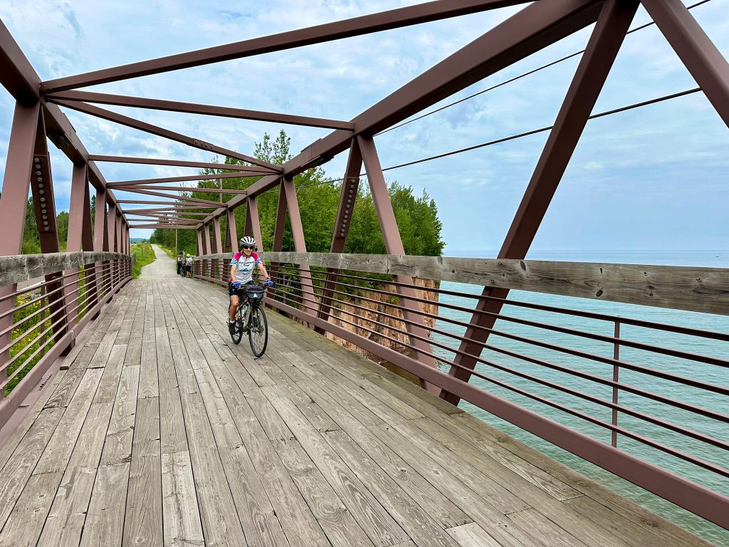 A cyclist riding over the wooden-planked truss bridge with Lake Superior in the background. 