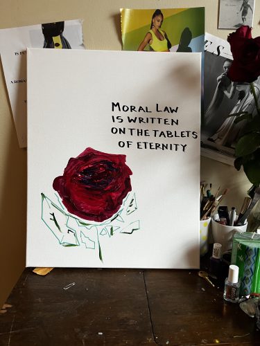 A painting of a rose with a quote in block text sits on the author's sewing table. She was using a cheap sewing table as a kitchen table in her studio apartment. Now she uses it to sew. At the time, she would obsessively paint roses as she worked through insomnia. The roses would have inscriptions in block letters. This inscription is a quote attributable to Lord Action, "Moral law is written on the fabric of eternity."