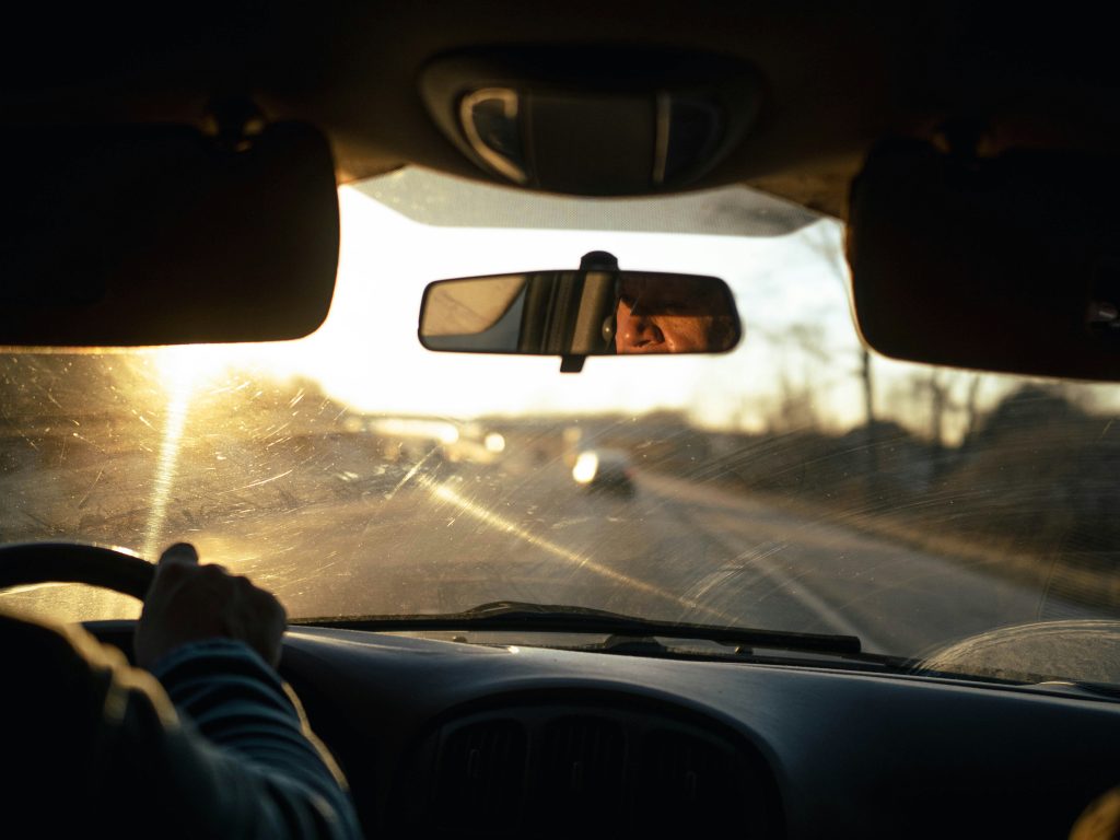 A view from the backseat of a driver, with his right hand visible on the wheel and his eyes, seen from the rearview mirror, staring intently at the road.