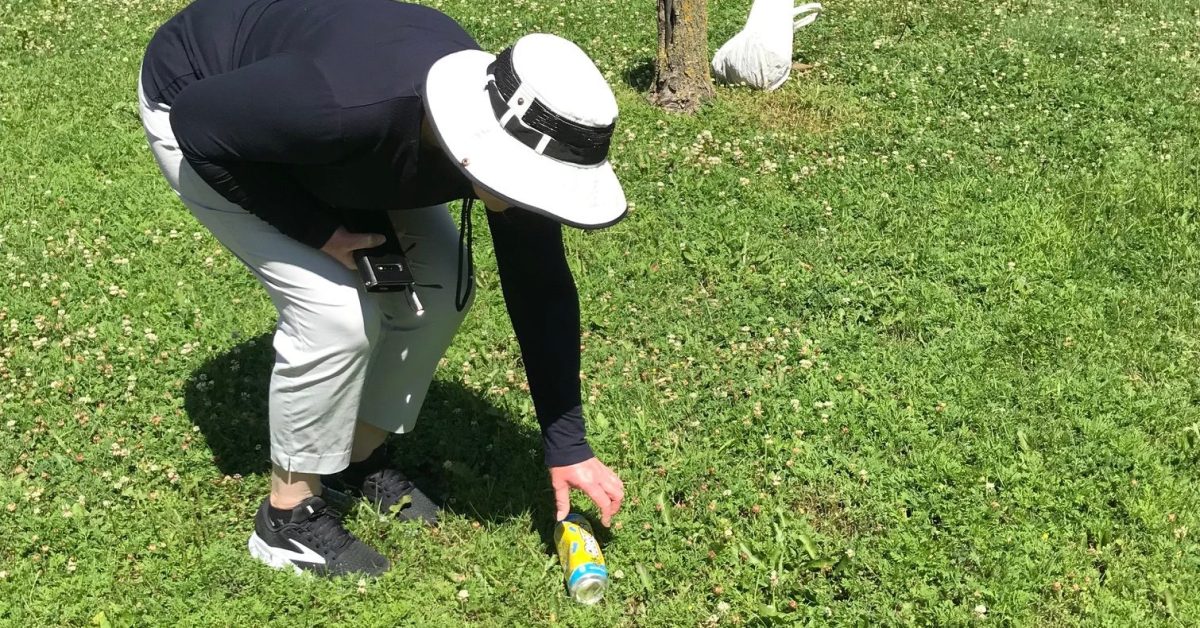 A person wearing a sun hat bends down to pick up trash on a lawn.