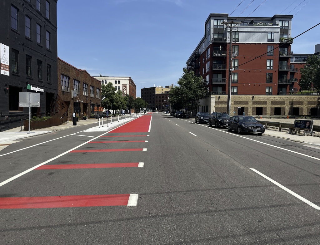 Look down a road with a separated bike lane on the left, concrete divider in the distance next, a red bus lane, and care lanes, with parked cars at the right