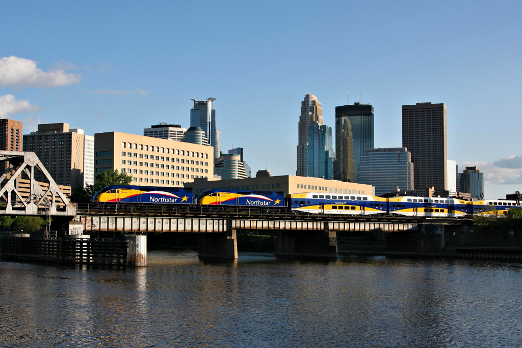 The Northstar Line train in blue, yellow and white with red accents crossing over the river against the Minneapolis skyline. 
