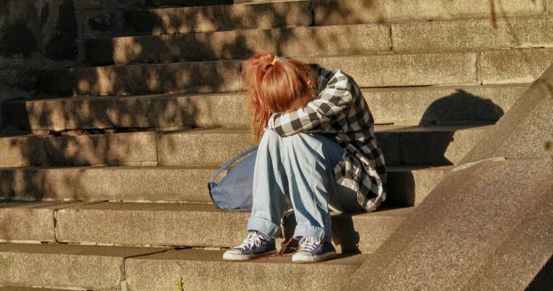 A woman with red hair and wearing jeans sits with her face on her knees on cement steps.