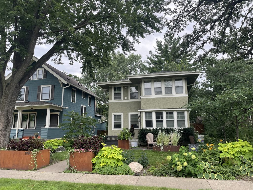 Two residential homes, the one on the left is a two-level house with blue siding, the one on the right a two-level home with taupe stucco. 