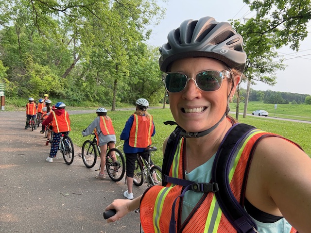 An adult woman wearing a bike helmet and safety vest poses for a selfie on her book while children line up behind her for a bicycling lesson.