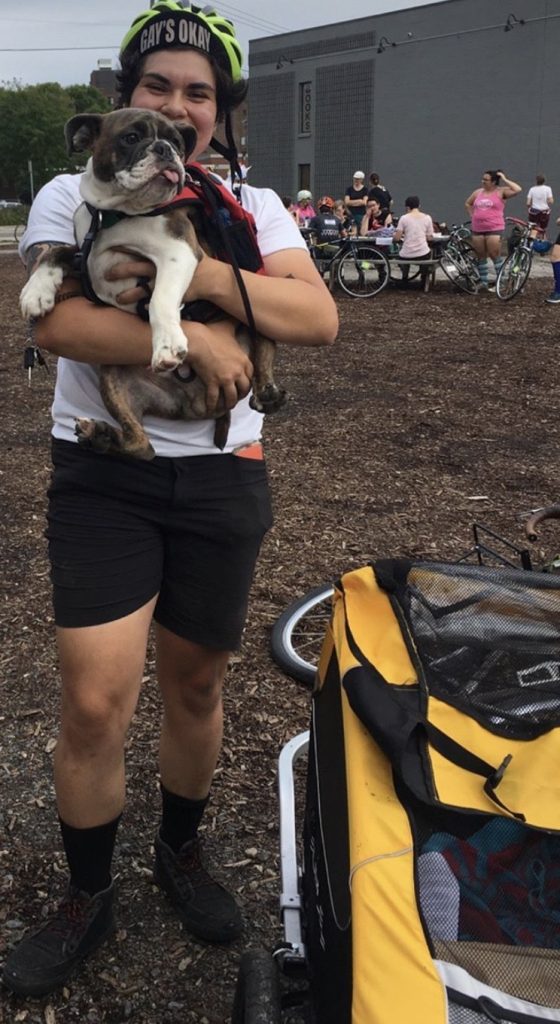 A smiling person is standing next to a bike trailer, holding a white-and-brown pitbull-looking dog. In the background, multiple people work on bikes.