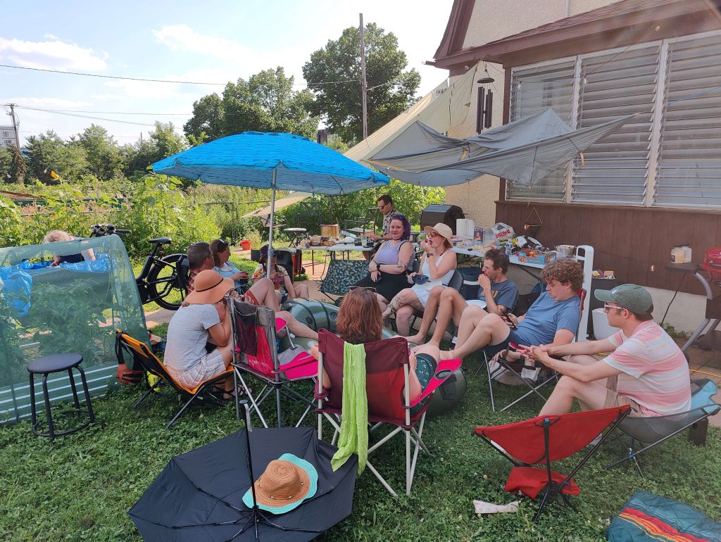 A group of friends sitting in a circle on lawn chairs in a backyard on a sunny day.