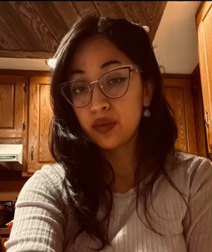 Pacha, a woman with slightly wavy, shoulder-length brown hair and glasses, looks at the camera with an unamused (yet badass) expression. She's wearing a white long-sleeve blouse and earrings.