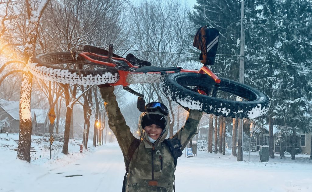 Cristina proudly holds their bike aloft on a snowy, tree-lined street.