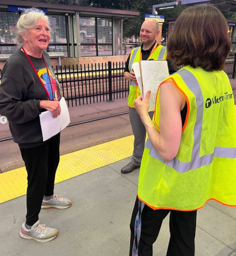 An older woman speaks with two Metro Transit workers in reflective vests as they conduct a rider survey at a light rail station.