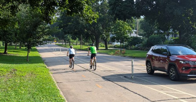 Two cyclists ride downhill in a separated bikeway while a car drives by in the other direction.