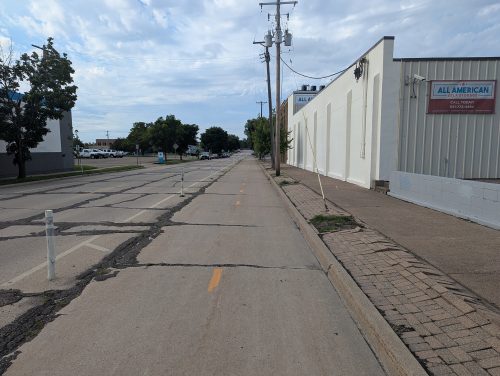A street with cracked pavement and a brick boulevard space that is also in poor condition.