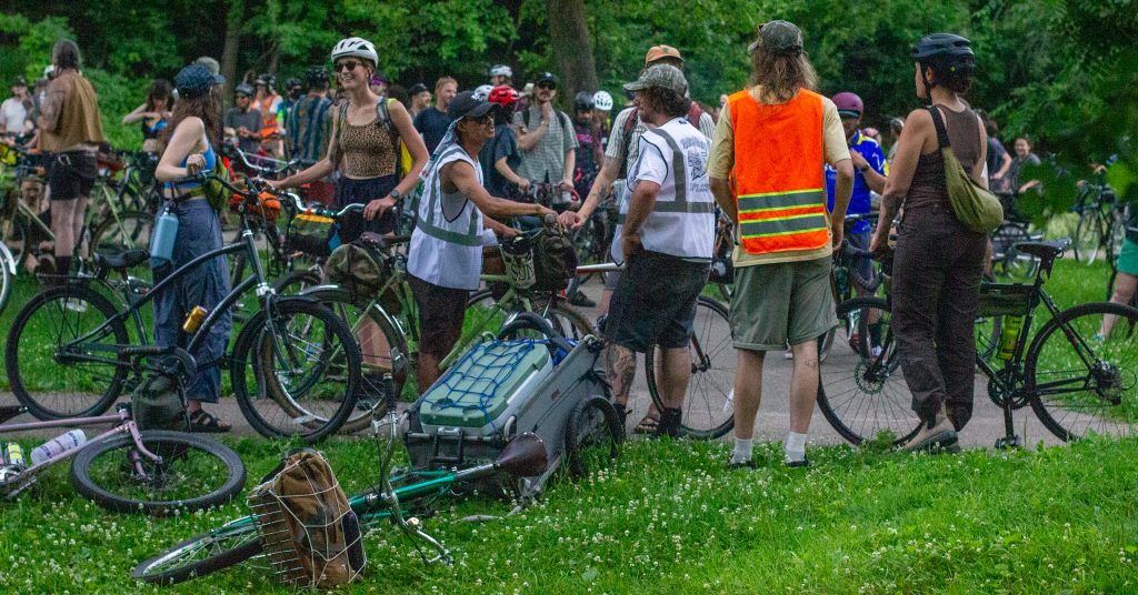 A group rests and socializes during a bike ride. There are smiling faces, handshakes, safety vests and bike trailers strewn about Minnehaha Creekside Park.