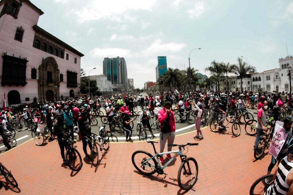 Hundreds of people on bicycles gather in a large city square for a group ride, surrounded by historic and modern buildings.