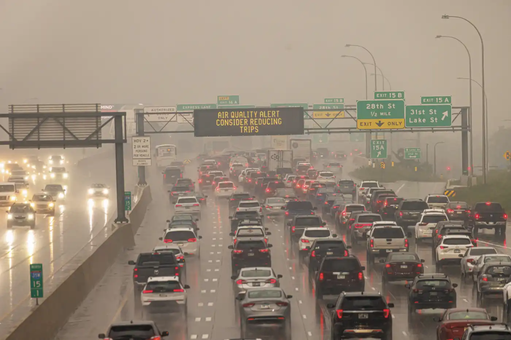 A congested highway in heavy haze and rain, with an overhead sign reading “Air Quality Alert: Consider Reducing Trips.”