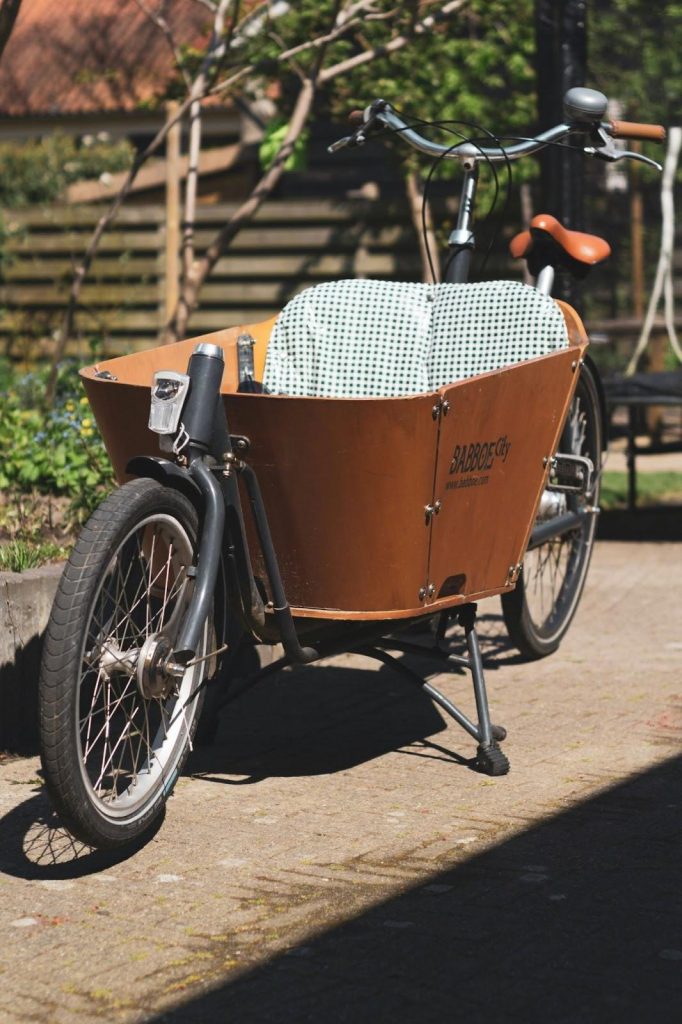 A front-loading cargo bicycle with a wooden box and patterned seat cover, parked outdoors on a sunny day.