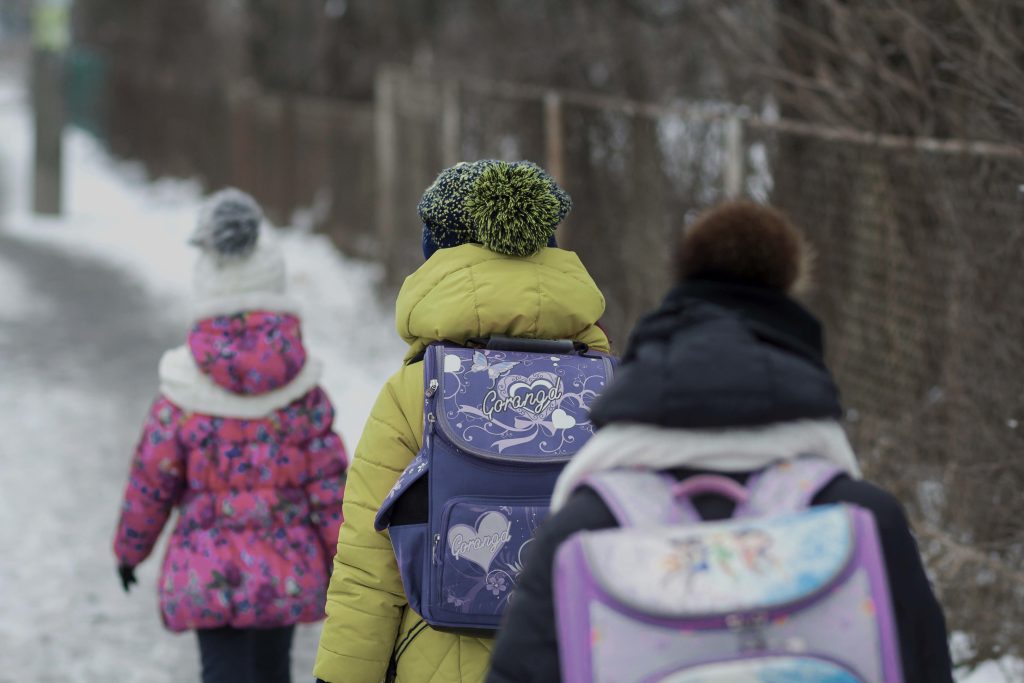 Three schoolchildren in winter jackets and hats are shown from behind walking on a snowy path.