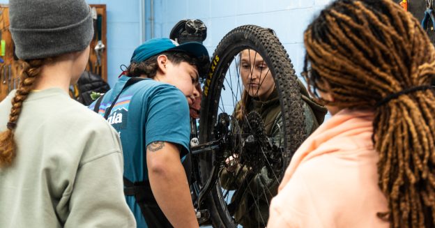 Three women surround a nonbinary person who is demonstrating how to fix a bike.