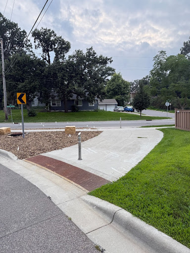 A residential street with a new concrete path curving around a landscaped median. Metal bollards block cars from entering the path, allowing only people walking or biking to pass through.