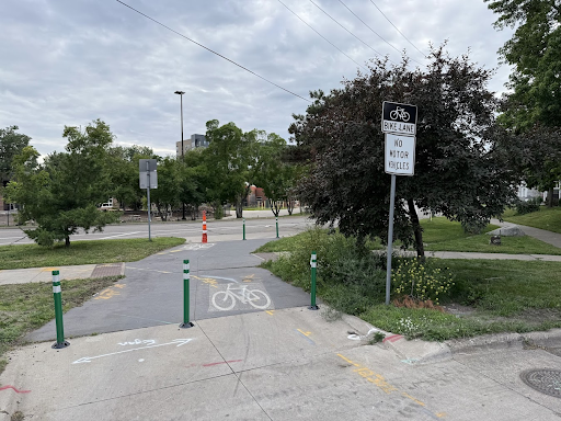 An entrance to a bike path marked with green bollards and a painted bicycle symbol on the pavement. A street sign reads “Bike Lane – No Motor Vehicles.” Trees and grass surround the path under a cloudy sky.