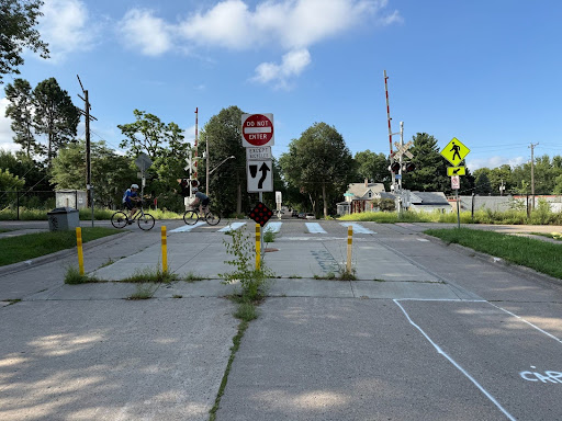 A street blocked by yellow bollards and a “Do Not Enter” sign, with a crosswalk leading to a railroad crossing. Two people on bikes ride through the opening, showing the space is reserved for walking and biking, not cars.
