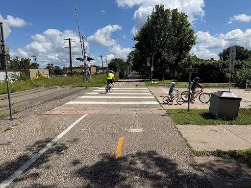 A bike trail crossing a street at a marked crosswalk near railroad tracks. A cyclist rides across while two children with bikes wait on the sidewalk, showing the trail is designed for people biking and walking rather than cars.