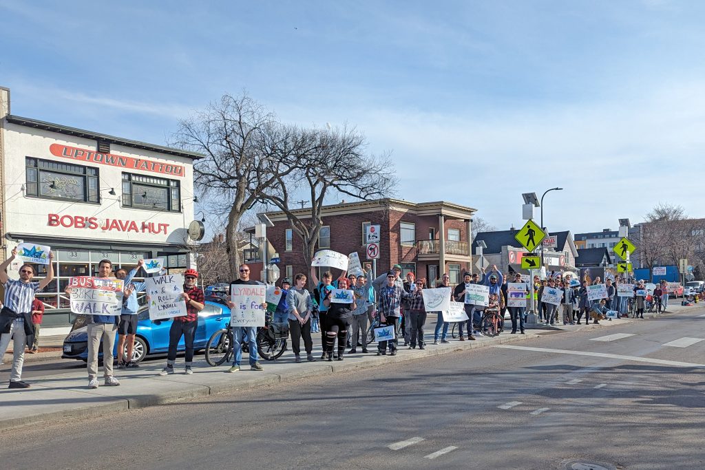 A large group of people stand on the median of Lyndale Ave S at W 27th St holding signs in support of transit, cycling, walking, safety, etc.