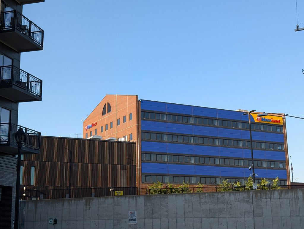 Building with MetroTransit logo on both its brick-veneer, gabled left face and its flat-roofed front face whcih alternates courses of windwos with courses of blue paneling, the top one giving way to yellow where the logo is