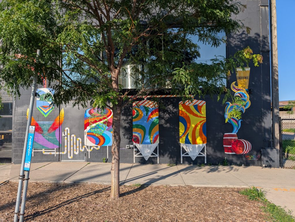 Grey cinderblock wall with colorful depiction of brewing process painted on it; the actual stainless-steel eqiuptment is just visible through a window, partially obscured by a tree