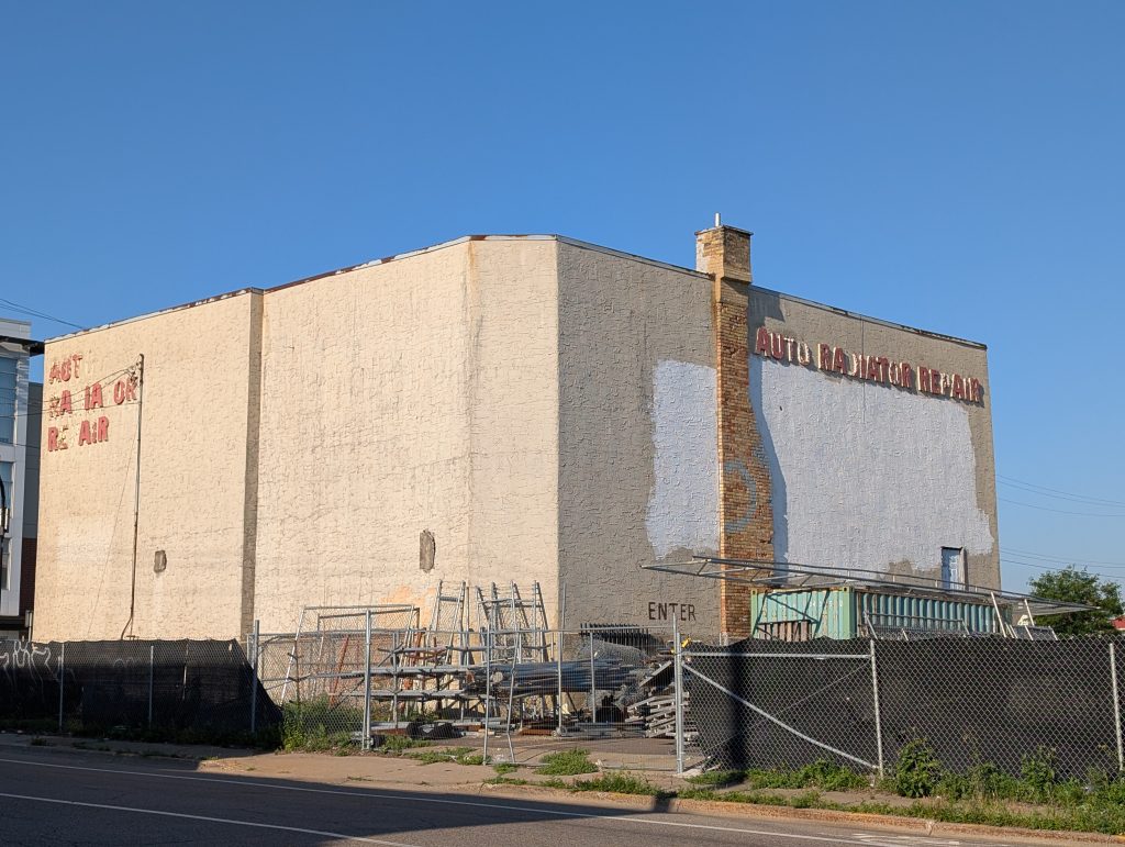 Boxy building with decaying lettering on two sides indicating AUTO RADIATOR REPAIR and a brick chimney