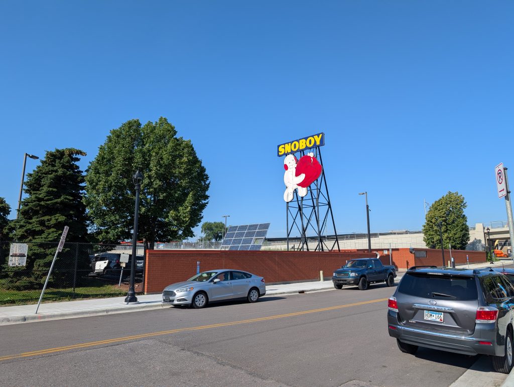 Street scene with prominent billboard for SNOBOY with a cartoonish character, apparently a snowman with arms and legs also formed from snowballs, wearing red earmuffs and holding a red bell pepper