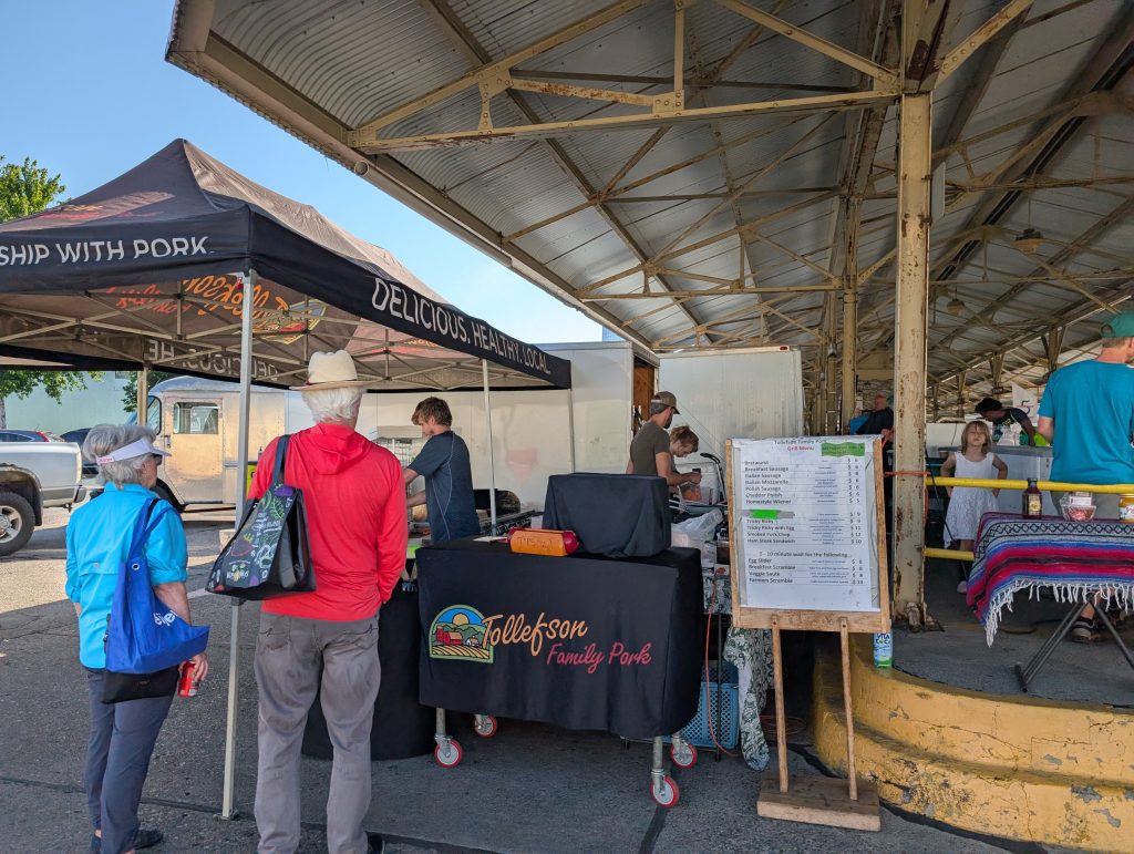 End view of one of the long raised-platform sheds comprising the farmers market; Tollefson Family Pork has a stand next to it