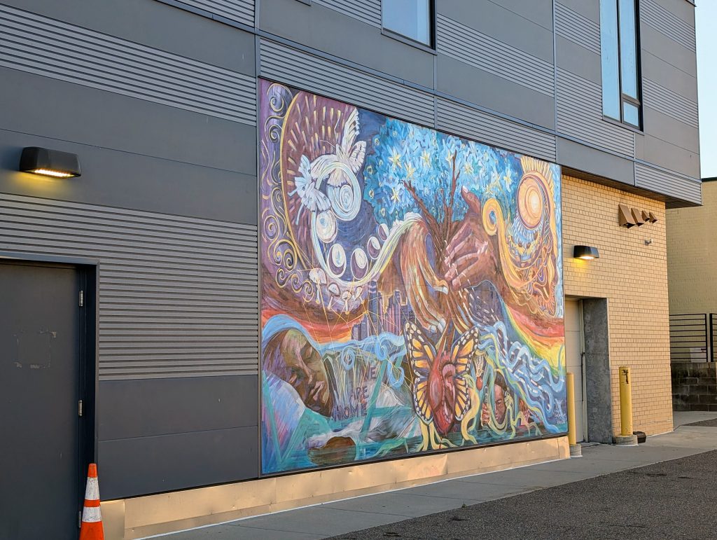 Mural featuring a figure at repose in its lower left corner accompanied by the inscription "we are home"