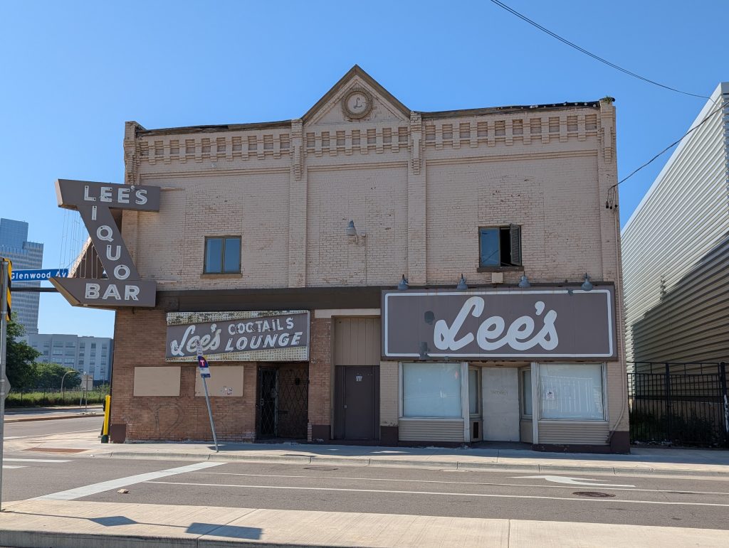Two-story tan building, Lee's Liquor Lounge, with signage of "Lee's," "Lee's COCKTAILS LOUNGE," and "LEE'S LIQUOR BAR," the last of these contrived to share the "L" between "LEE'S" and "LIQUOR" and the "R" between "LIQUOR" and "BAR"; there's also an "L" rosette in the gable