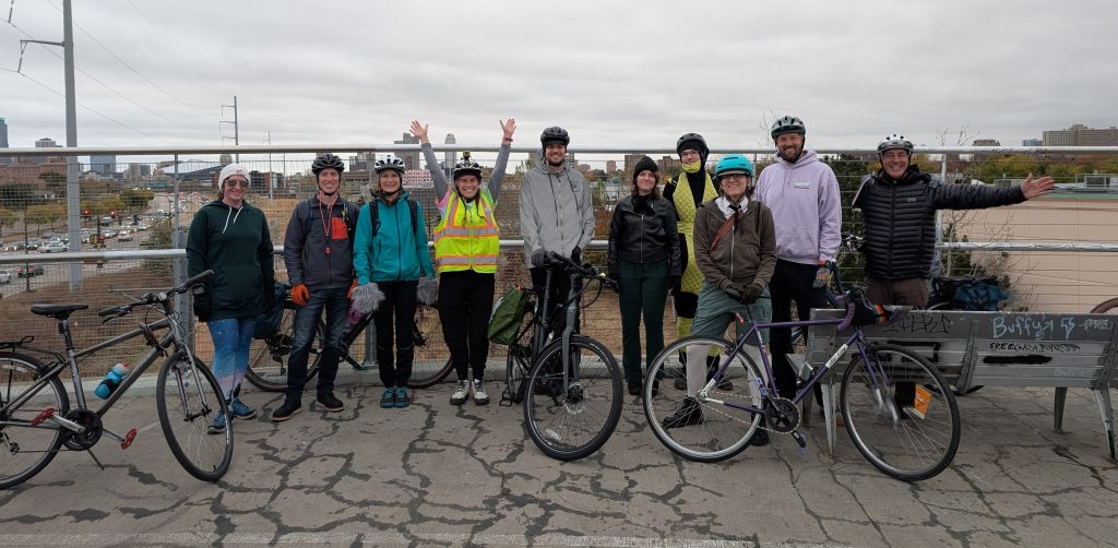 A group of ten cyclists wearing helmets and cold-weather gear stand together smiling on a bridge, with their bikes in front of them. A city skyline and busy highway are visible in the background under a cloudy sky.