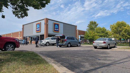 Exterior view of an Aldi grocery store. Several cars are parked in front, with people walking toward the entrance under a partly cloudy sky.
