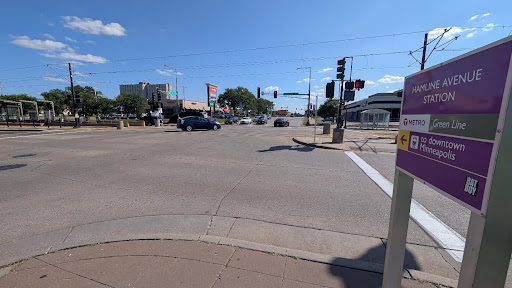 Street view from Hamline Avenue Green Line Station, looking across a wide intersection toward shops and traffic lights. A station sign is in the foreground, with directions toward downtown Minneapolis.