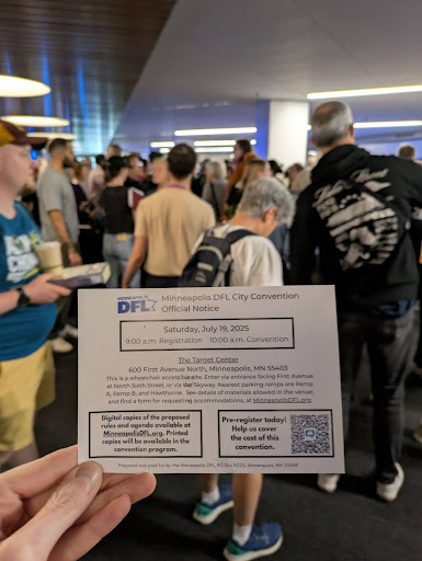 A person holds up an official Minneapolis DFL City Convention notice card while standing in a crowded lobby at the Target Center. The card lists the convention date, July 19, 2025, and details about registration and location. In the background, people wait in line and talk in small groups under bright overhead lights.