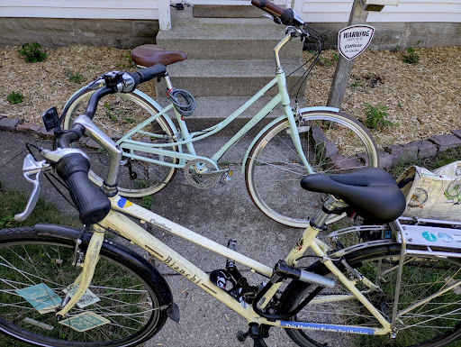 Two bicycles are parked near a small set of concrete steps outside a house, one pale yellow and one light blue. A “Warning: Security Camera” sign is visible beside them.