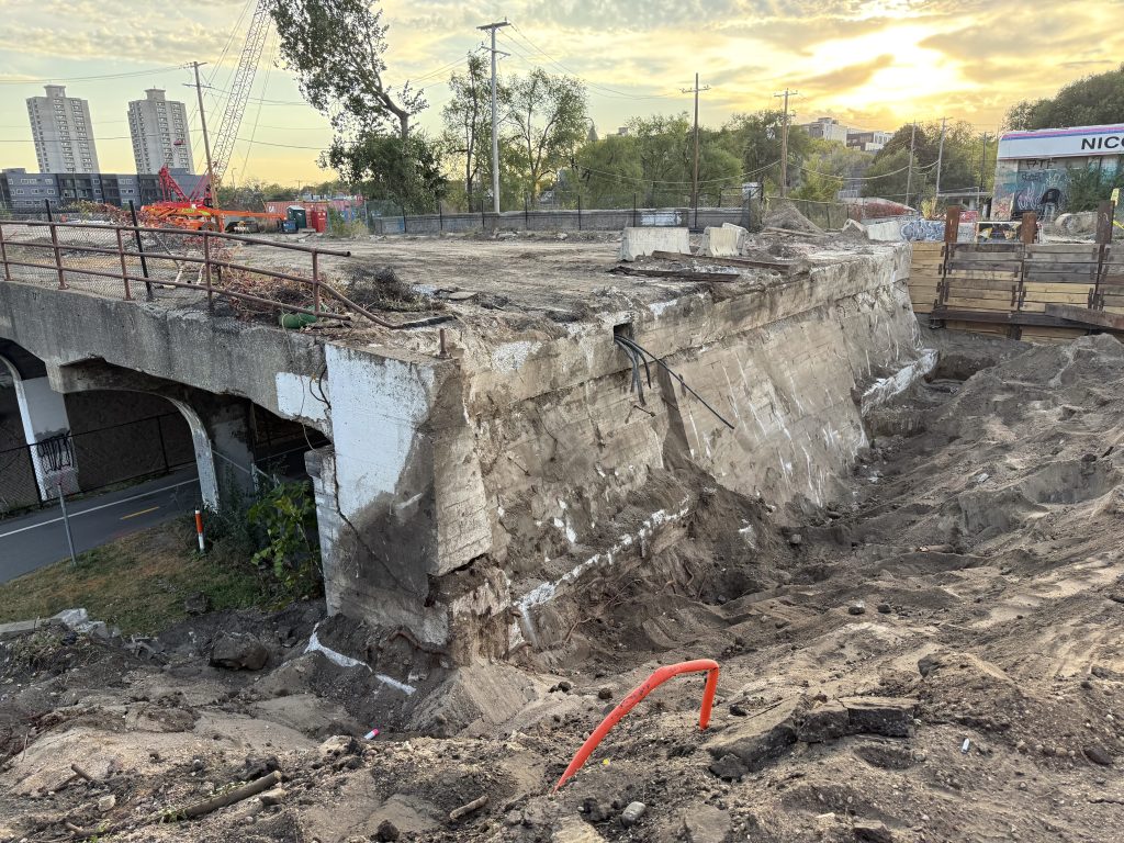 Looking southwest from the northeast corner of the Nicollet Ave bridge, most of the north abutment is exposed, with utilities and cut streetcar rails gaining off the edge.