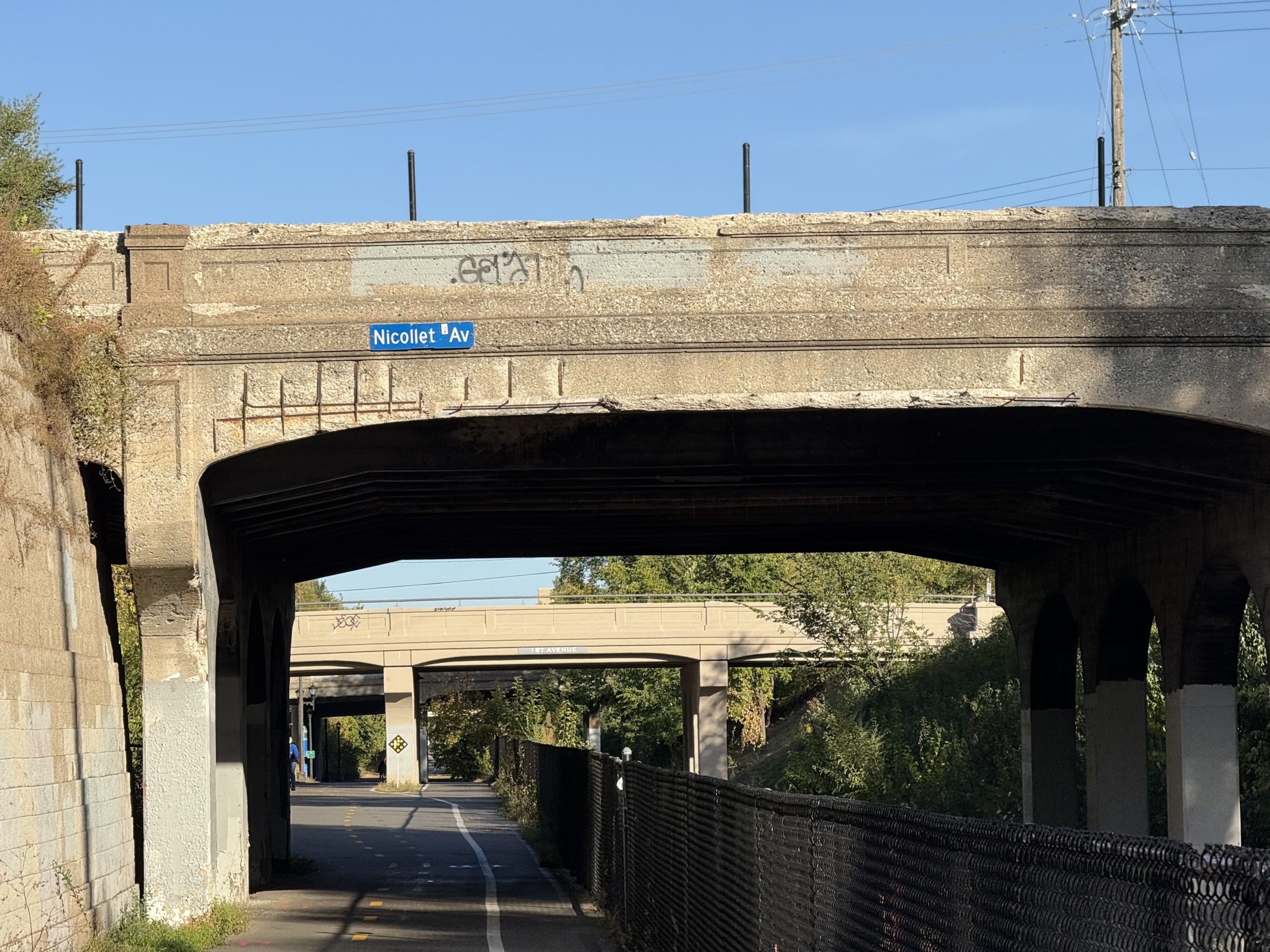 Side profile of Nicollet Avenue bridge with the Midtown Greenway path going under, and a street sign saying Nicollet Ave mounted on the side of the bridge.