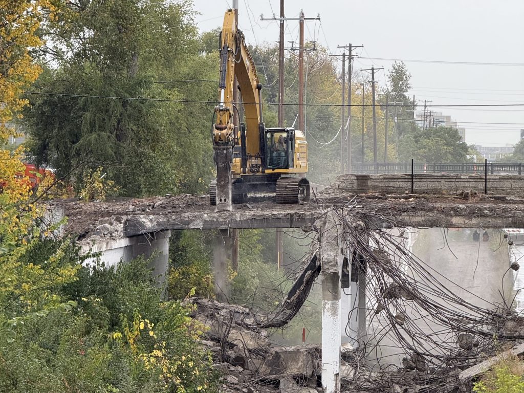 Side profile of the Nicollet Ave bridge looking to the west, an excavator sits on the bridge while breaking up the concrete next to it. Bands of concrete and rebar hang off of the piers.