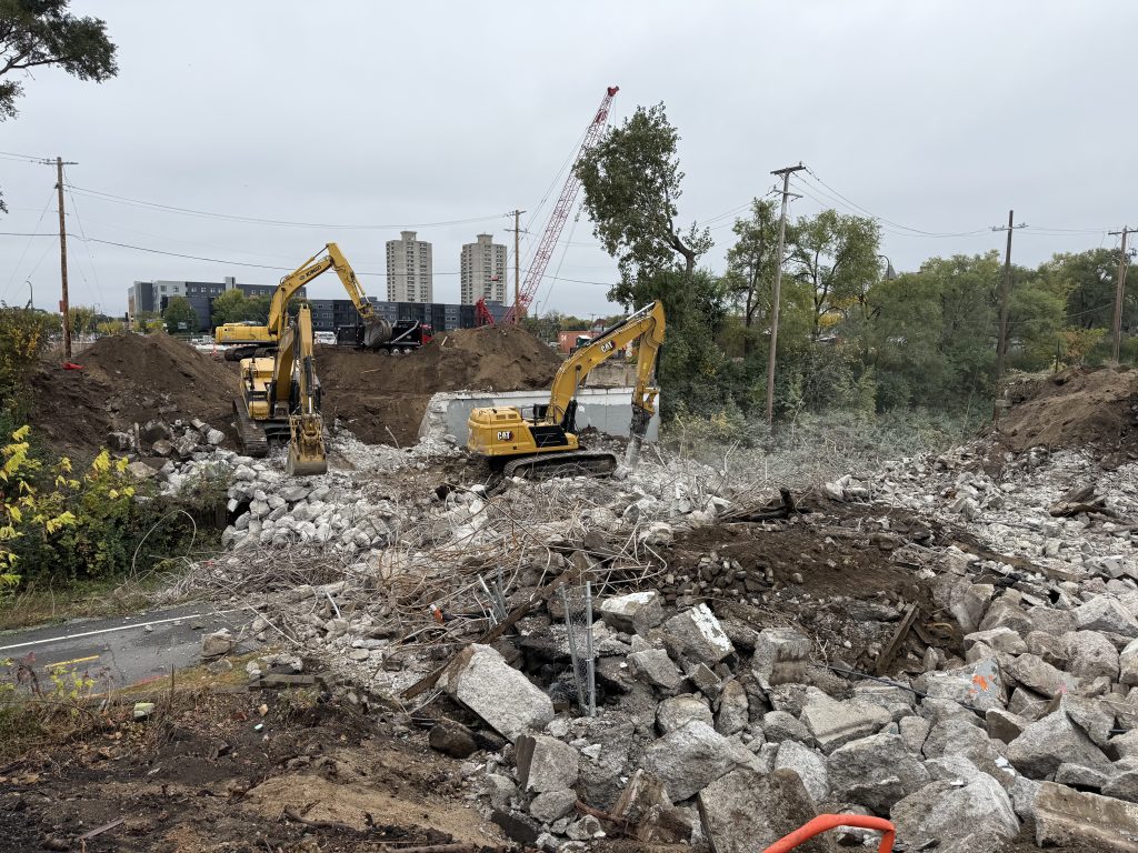 Three excavators work on breaking up, sorting, and moving a large pile of concrete and rebar where the Nicollet Ave bridge once stood. The southern abutment is roughly 60% still intact.