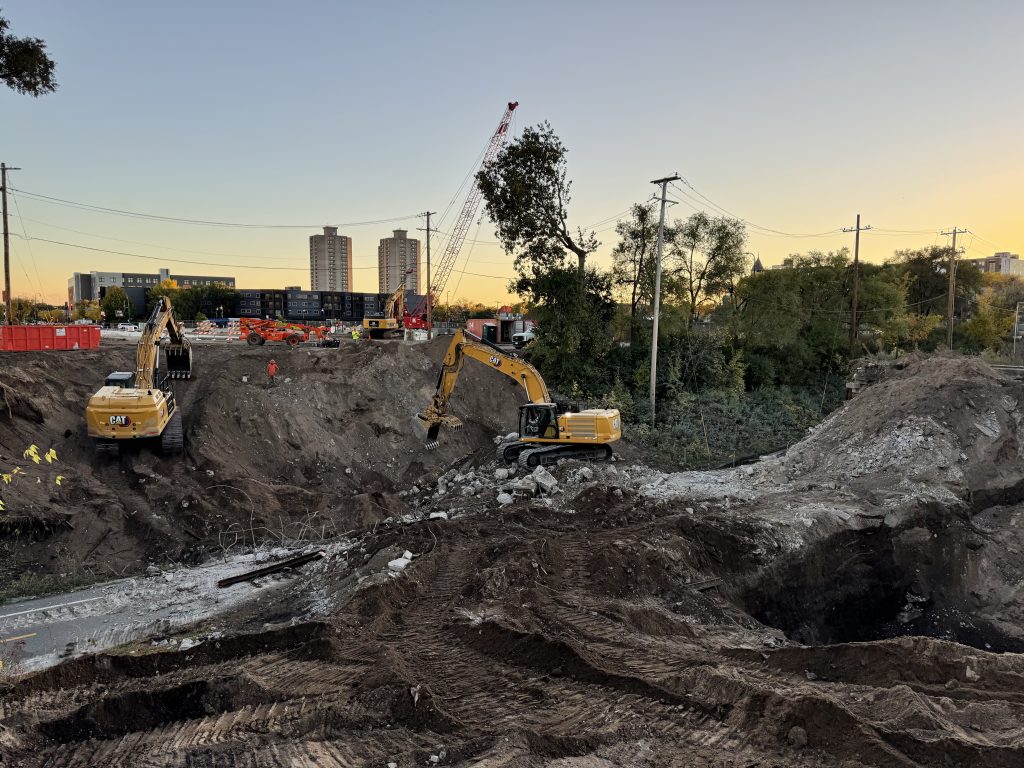 Some concrete, but mostly dirt covers the Midtown Greenway trail where the Nicollet Ave bridge once stood. Three excavators are working and a crane sits in the background.