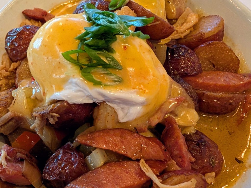 layered food, listed from top to bottom: garnish of diagonally sliced scallions, spicy hollandaise, poached  egg, hash, sauce